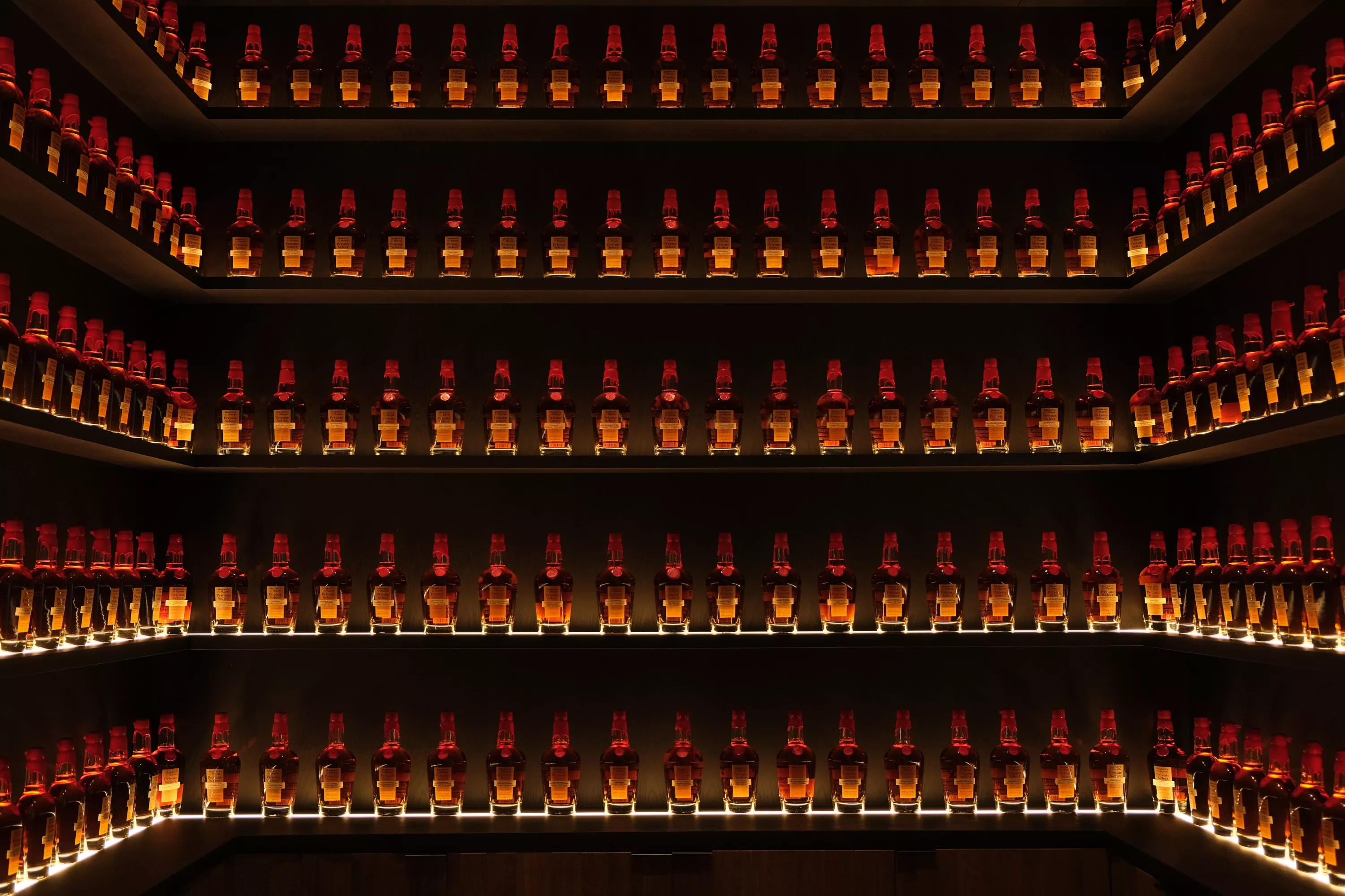 Three walls with four rows of glass shelves against a dark backdrop holding rows of Maker's Mark Limited Release series handles.