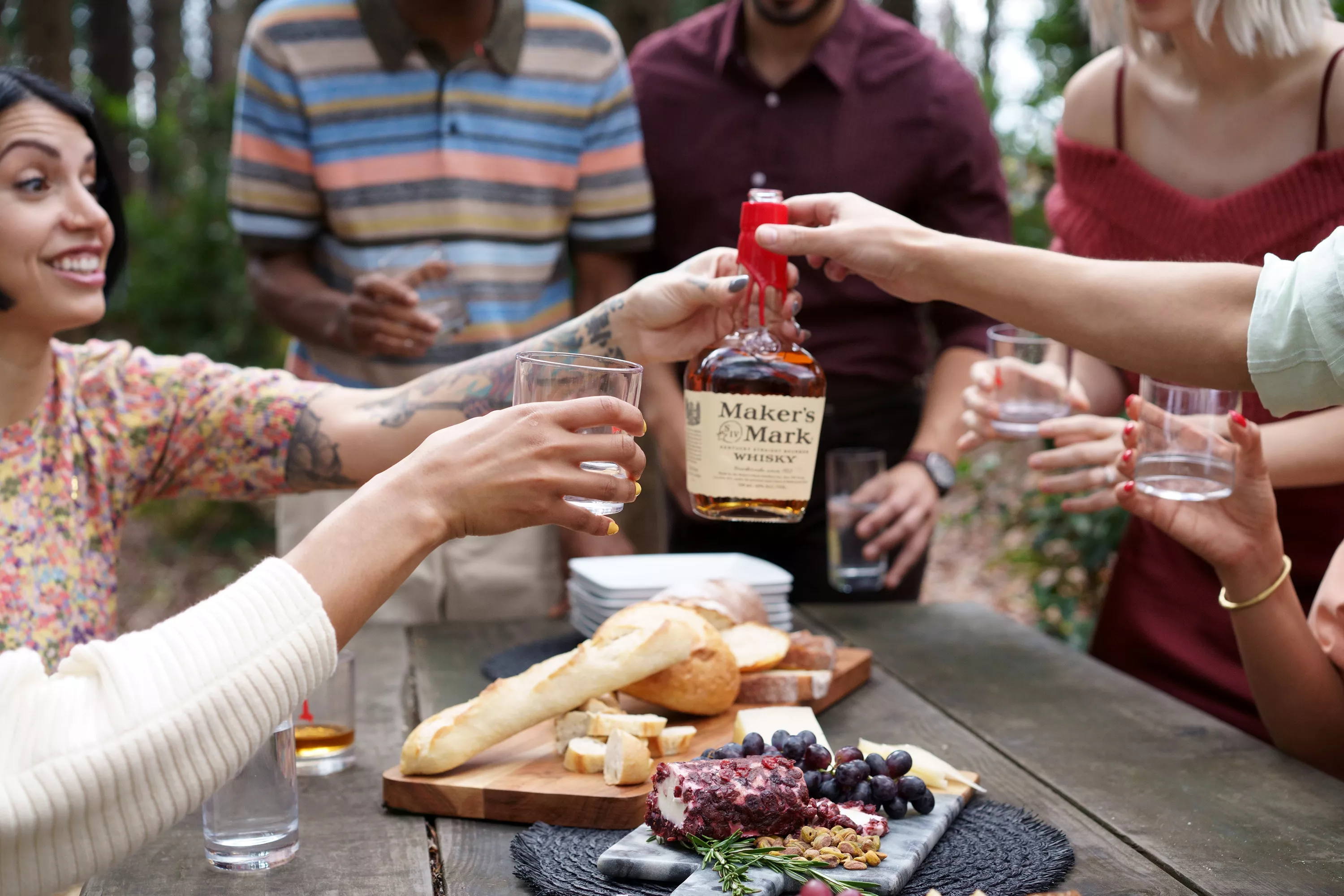 A group of friends outdoors toasting with Maker's Mark bourbon whisky around a picnic table with a charcuterie board set out.