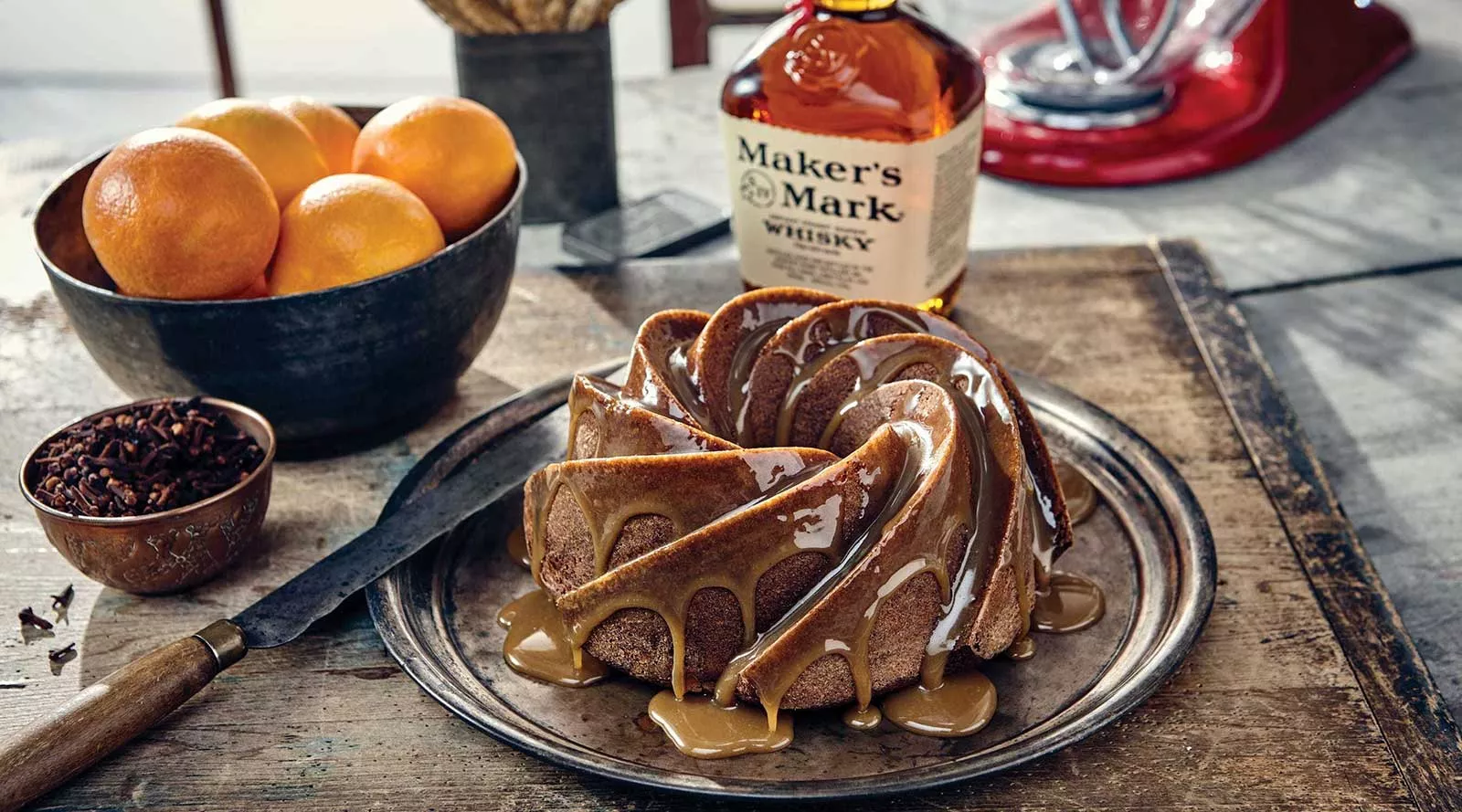 A glazed bundt cake sits on a round baking sheet, next to a bowl of fresh oranges, spices, and a bottle of Maker's Mark.